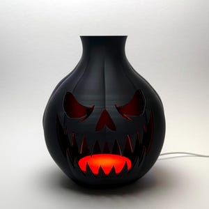 Puede incluir: L&aacute;mpara decorativa negra con tem&aacute;tica de Halloween con forma de linterna de calabaza. La l&aacute;mpara tiene una cara tallada con ojos y boca rojos brillantes. La l&aacute;mpara est&aacute; enchufada y emite una luz naranja c&aacute;lida.