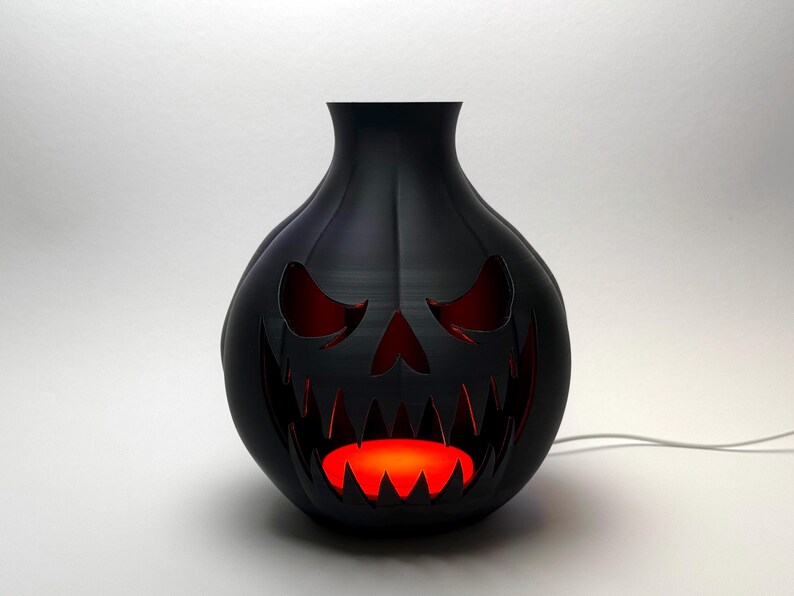 Puede incluir: L&aacute;mpara decorativa negra con tem&aacute;tica de Halloween con forma de linterna de calabaza. La l&aacute;mpara tiene una cara tallada con ojos y boca rojos brillantes. La l&aacute;mpara est&aacute; enchufada y emite una luz naranja c&aacute;lida.