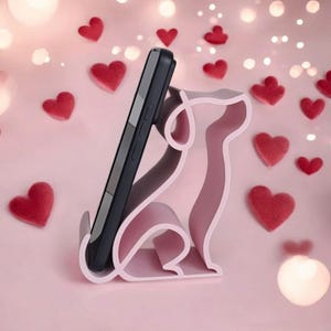 May include: A light pink dog-shaped phone stand holding a black smartphone. The stand is surrounded by red heart-shaped decorations on a pink background. The image has a soft, romantic feel.