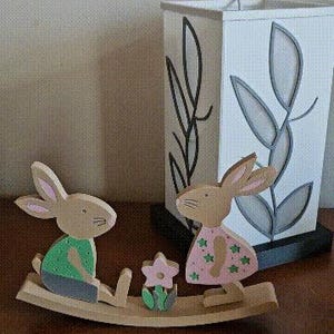 May include: Wooden Easter bunny figurines on a wooden rocker with a flower. One bunny wears a green shirt and gray shorts, the other a pink dress with green stars. A white lamp with a black leaf design is in the background.