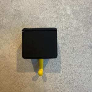 May include: A black, rectangular wall hook with a yellow hook. The hook is mounted on a light gray textured wall. The hook is designed to hold items and is a simple, modern design.
