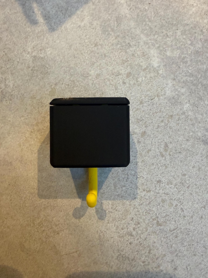 May include: A black, rectangular wall hook with a yellow hook. The hook is mounted on a light gray textured wall. The hook is designed to hold items and is a simple, modern design.