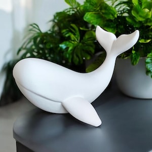 May include: A white ceramic whale figurine with a smooth finish. The whale has a curved body, a tail, and a fin. The figurine is displayed on a dark gray surface, with green plants in the background.