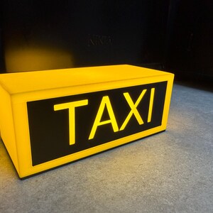 May include: A rectangular yellow taxi sign with the word "TAXI" in bold, yellow letters against a black background. The sign is illuminated, suggesting it is lit from within. The sign is sitting on a gray surface.