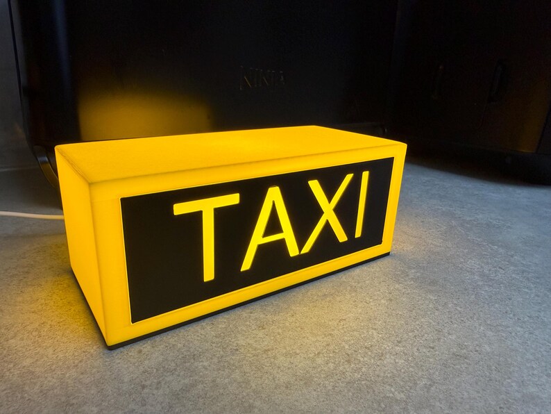 May include: A rectangular yellow taxi sign with the word "TAXI" in bold, yellow letters against a black background. The sign is illuminated, suggesting it is lit from within. The sign is sitting on a gray surface.