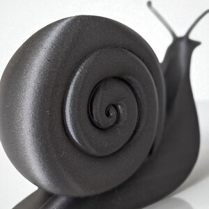May include: A black, three-dimensional snail figurine with a textured surface. The snail's shell features a spiral design, and its body includes antennae and a base. The figurine is set against a white background.