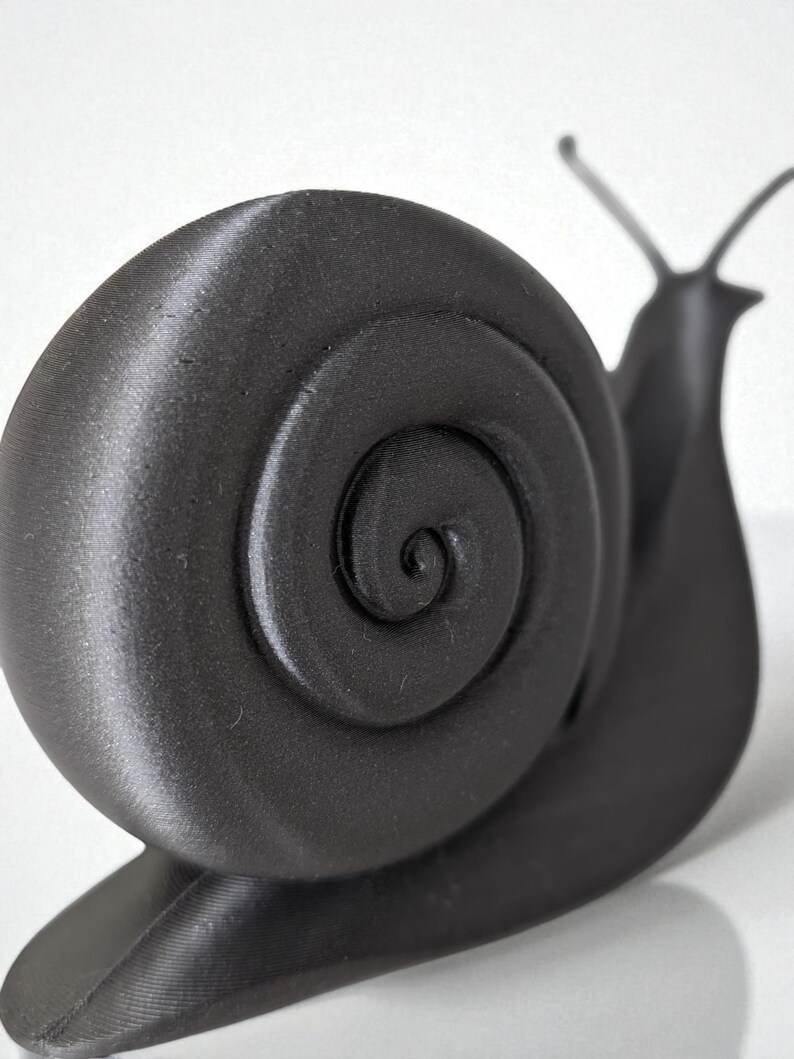 May include: A black, three-dimensional snail figurine with a textured surface. The snail's shell features a spiral design, and its body includes antennae and a base. The figurine is set against a white background.