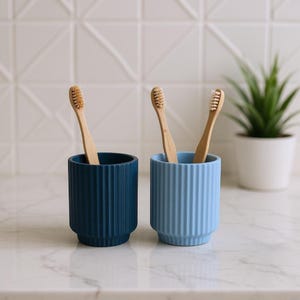 May include: Two ceramic toothbrush holders, one in navy blue and the other in light blue, each holding wooden toothbrushes. The holders have a ribbed design. A small potted plant is in the background, adding a touch of greenery to the scene.