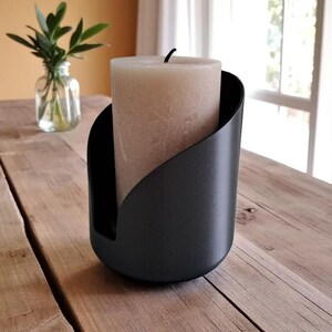 May include: A black candle holder with a curved design holds a beige pillar candle. The candle holder is made of a matte black material and sits on a wooden surface. A small glass vase with green leaves is in the background.