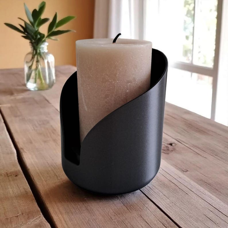 May include: A black candle holder with a curved design holds a beige pillar candle. The candle holder is made of a matte black material and sits on a wooden surface. A small glass vase with green leaves is in the background.