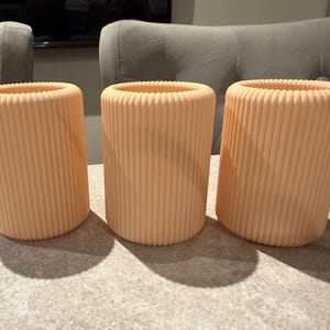 May include: Three peach-colored cylindrical vases with vertical ribbed detailing. The vases are arranged on a light gray surface, casting shadows. The open tops of the vases are visible, suggesting they are designed to hold items. The background includes a gray chair.