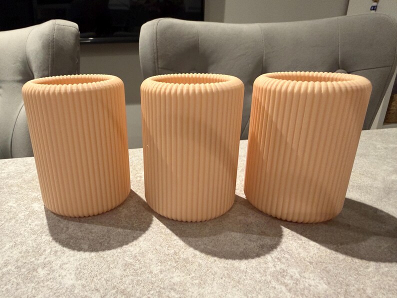 May include: Three peach-colored cylindrical vases with vertical ribbed detailing. The vases are arranged on a light gray surface, casting shadows. The open tops of the vases are visible, suggesting they are designed to hold items. The background includes a gray chair.