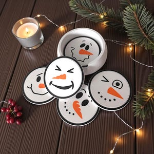 May include: A set of white snowman-themed coasters with black outlines and orange carrot noses. The coasters have different facial expressions and are displayed with a holder, a lit candle, and festive greenery. The coasters are approximately 4 inches in diameter.