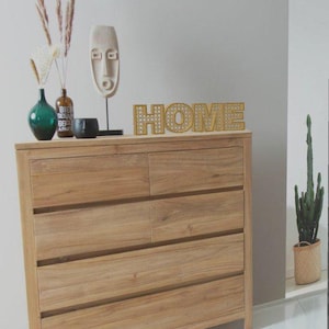 May include: A light-colored wooden dresser with six drawers, topped with decorative items. A gold-colored "HOME" sign sits on top, along with a green vase, a small black bowl, and a wooden mask sculpture. A cactus in a woven basket is visible.