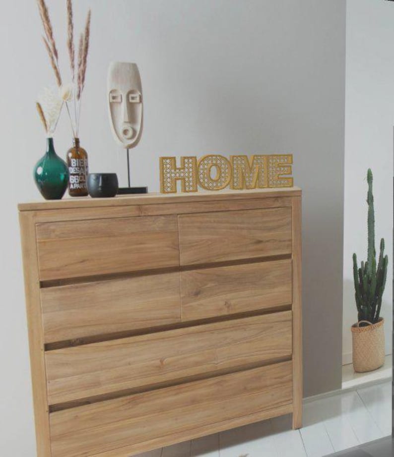 May include: A light-colored wooden dresser with six drawers, topped with decorative items. A gold-colored "HOME" sign sits on top, along with a green vase, a small black bowl, and a wooden mask sculpture. A cactus in a woven basket is visible.