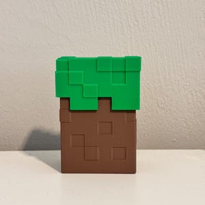 May include: A Minecraft-themed block, featuring a brown, pixelated base topped with green pixelated grass. The block is a solid, three-dimensional object, likely a decorative item or toy.