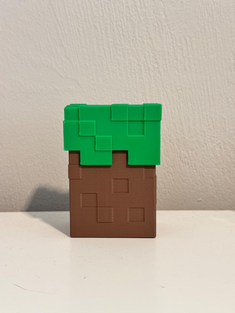 May include: A Minecraft-themed block, featuring a brown, pixelated base topped with green pixelated grass. The block is a solid, three-dimensional object, likely a decorative item or toy.