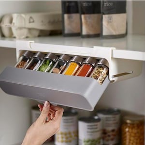 May include: A gray pull-out spice rack with clear glass jars filled with various spices. The rack is mounted inside a white cabinet. The jars have silver lids and are arranged in a row. The rack is being pulled out by a hand.