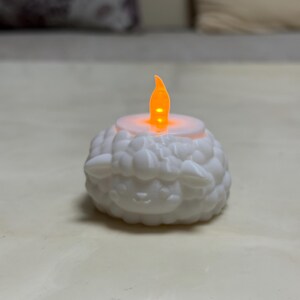 May include: A white, battery-operated LED candle shaped like a sheep. The candle has a flickering orange flame and is designed to resemble a sheep's wool. The candle is a decorative item.
