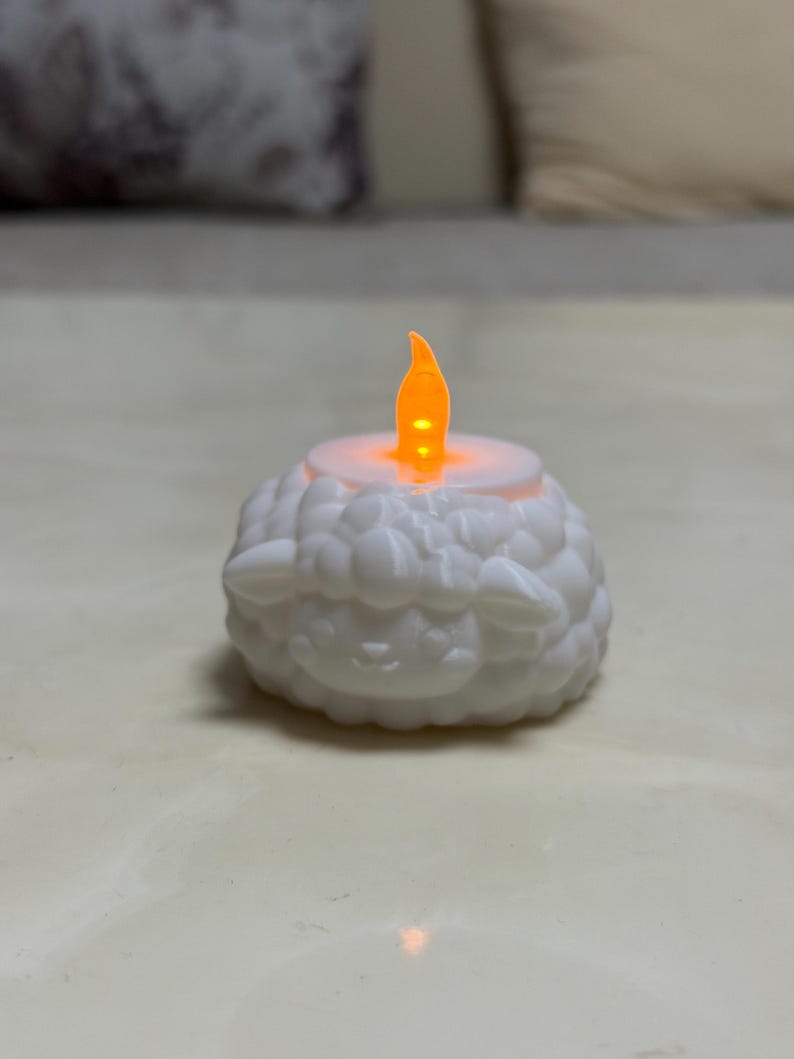 May include: A white, battery-operated LED candle shaped like a sheep. The candle has a flickering orange flame and is designed to resemble a sheep's wool. The candle is a decorative item.