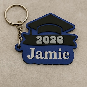 K&ouml;nnte beinhalten: Ein blauer Schl&uuml;sselanh&auml;nger zum Thema Abschluss mit dem Namen "Jamie" und dem Jahr "2026" in wei&szlig;en Buchstaben. Der Schl&uuml;sselanh&auml;nger hat ein Design einer Abschlusskappe in Schwarz und Blau, befestigt an einem silbernen Schl&uuml;sselring und einer Kette.