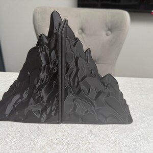 May include: Black mountain-shaped bookends. The bookends are made of a solid, dark material and feature a detailed, textured surface resembling a rocky mountain range. The bookends are designed to hold books upright on a desk or shelf.