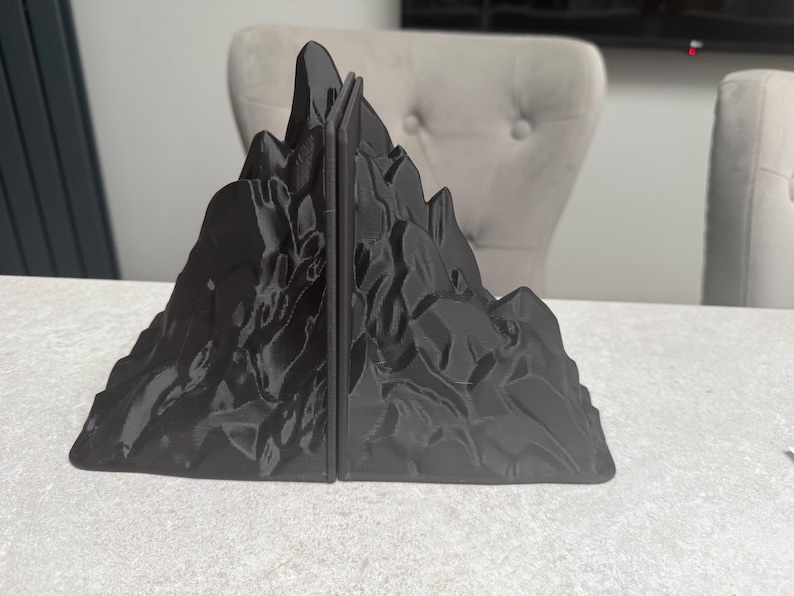 May include: Black mountain-shaped bookends. The bookends are made of a solid, dark material and feature a detailed, textured surface resembling a rocky mountain range. The bookends are designed to hold books upright on a desk or shelf.