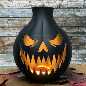 Puede incluir: Decoraci&oacute;n de Halloween con forma de calabaza negra con una cara tallada. Los ojos, la nariz y la boca est&aacute;n recortados, revelando un brillo naranja desde el interior. La decoraci&oacute;n est&aacute; sobre un lecho de hojas de oto&ntilde;o.