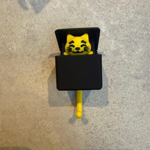 May include: A yellow cat-shaped hook with a black box and a yellow tail. The cat has a smiling face and is emerging from the black box. The hook is designed to hang on a wall or other surface.