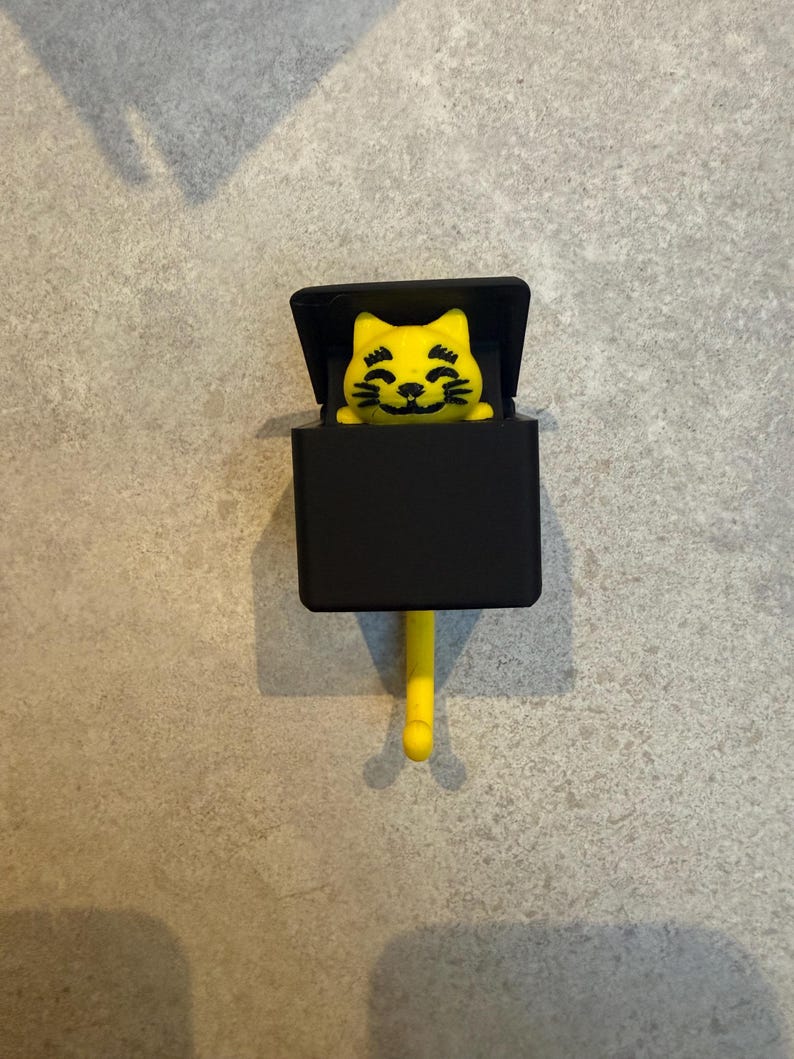 May include: A yellow cat-shaped hook with a black box and a yellow tail. The cat has a smiling face and is emerging from the black box. The hook is designed to hang on a wall or other surface.
