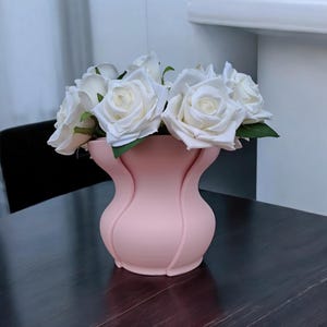 Puede incluir: Un jarr&oacute;n rosa con un dise&ntilde;o curvo &uacute;nico contiene un ramo de rosas blancas. Las rosas tienen hojas verdes y est&aacute;n en plena floraci&oacute;n. El jarr&oacute;n est&aacute; hecho de un material liso y descansa sobre una superficie de madera oscura.