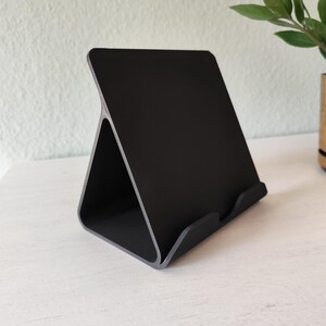 May include: A black tablet stand with a triangular base and a slightly angled back. The stand is made of a matte black material with a gray trim, designed to hold a tablet or similar device. The stand is on a white surface.
