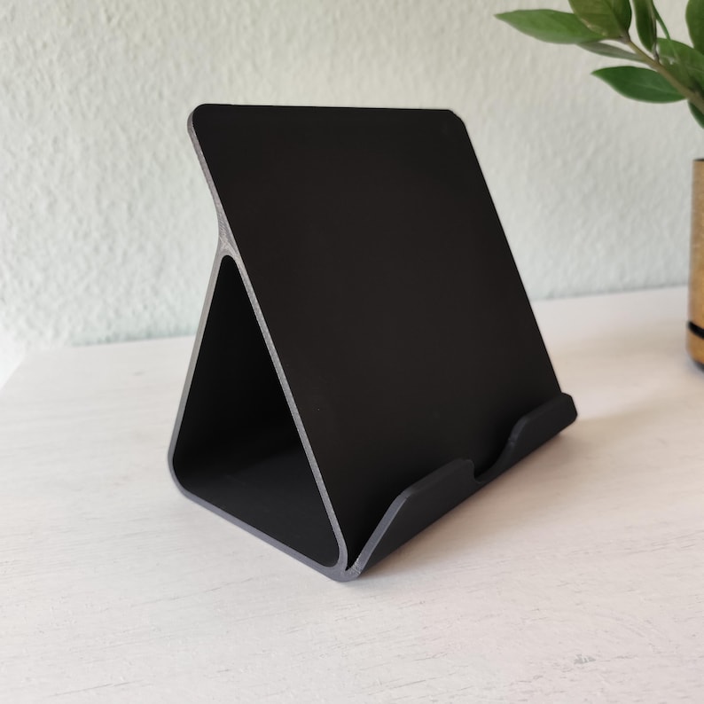 May include: A black tablet stand with a triangular base and a slightly angled back. The stand is made of a matte black material with a gray trim, designed to hold a tablet or similar device. The stand is on a white surface.
