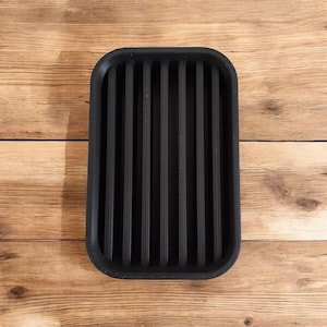 May include: A rectangular black soap dish with vertical ridges. The dish is made of a solid material and has rounded corners. The background is a light brown wooden surface.