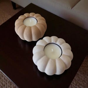 May include: Two decorative candle holders with a pumpkin-like design. One is a light beige color, and the other is white. Each holder has a circular opening to hold a white candle. The holders are on a dark brown surface.
