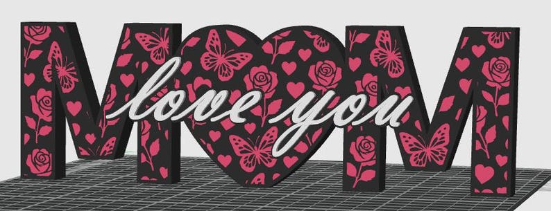 May include: A decorative sign spelling "MOM" with a heart in the center. The letters and heart are black with pink butterfly, rose, and heart patterns. The words "love you" are written in white across the heart.