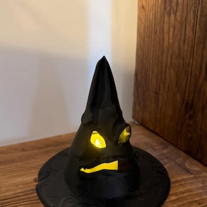 Op de afbeelding: Een zwarte decoratieve heksenhoed met een heldergeel licht van binnen. De hoed heeft een brede rand en een puntige top, en staat op een houten oppervlak. Een Halloween decoratie.