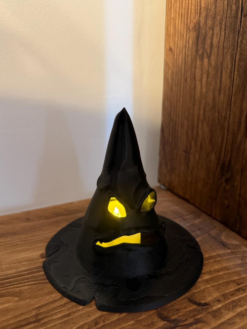 Op de afbeelding: Een zwarte decoratieve heksenhoed met een heldergeel licht van binnen. De hoed heeft een brede rand en een puntige top, en staat op een houten oppervlak. Een Halloween decoratie.
