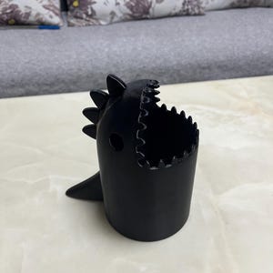 May include: A black, dinosaur-shaped container with a wide open mouth and a small fin. The container has a cylindrical body and is sitting on a light-colored surface. The dinosaur has a spiky back.