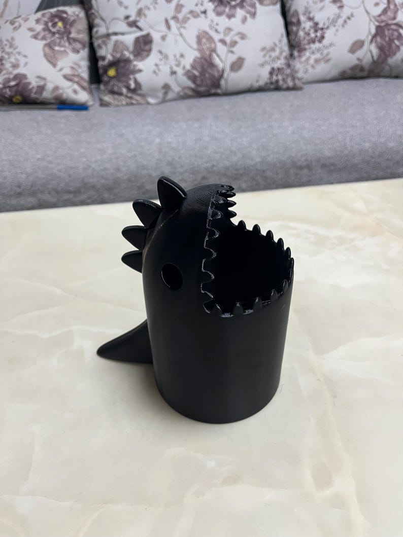 May include: A black, dinosaur-shaped container with a wide open mouth and a small fin. The container has a cylindrical body and is sitting on a light-colored surface. The dinosaur has a spiky back.