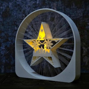 May include: A white star-shaped decorative light with a cut-out snowflake design and a reindeer silhouette. The star is encased in a white circular frame with radiating lines, creating a festive, illuminated display.
