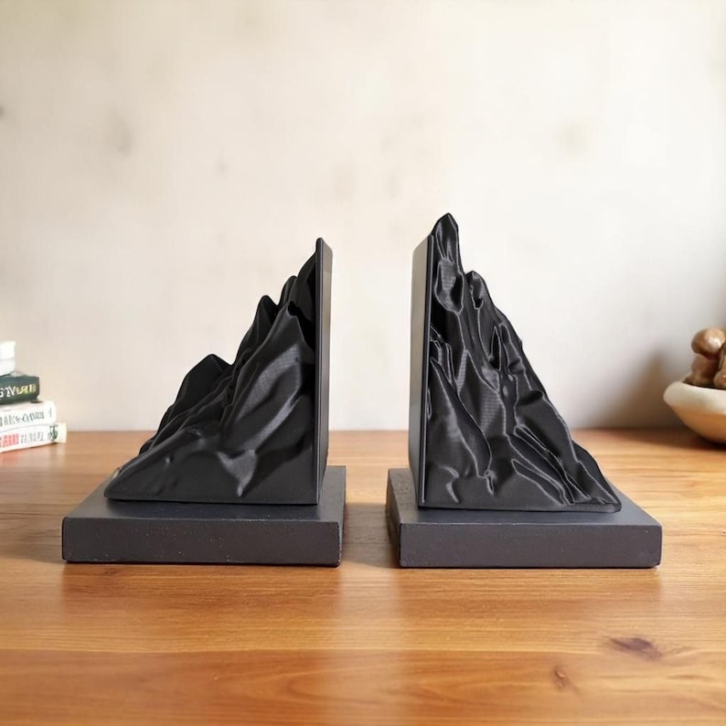 May include: Black mountain-shaped bookends. Each bookend features a detailed, textured mountain range design atop a rectangular base. The bookends are made of a solid, dark material and are designed to hold books upright. The bookends are approximately 6 inches tall.
