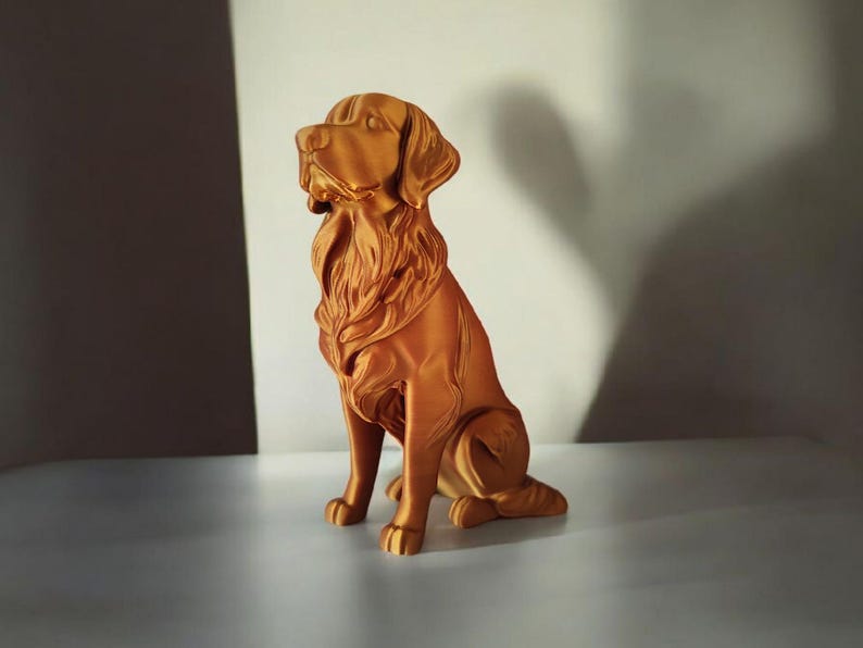 May include: A golden-colored dog statue in a sitting position. The statue has detailed fur and a regal posture, with its head held high. The sculpture is made of a solid material and is displayed on a white surface.