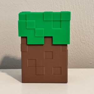 May include: A Minecraft-themed block, featuring a brown, textured base with green, pixelated top. The block is a cube shape, with a simple, geometric design. The color scheme is brown and green.