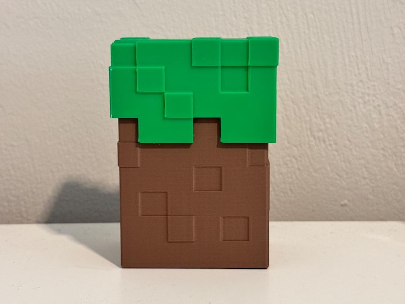 May include: A Minecraft-themed block, featuring a brown, textured base with green, pixelated top. The block is a cube shape, with a simple, geometric design. The color scheme is brown and green.