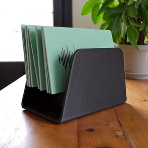 May include: A black desk organizer holding several light green cards with a black graphic design. The organizer is made of a textured material and sits on a wooden surface. A potted plant is in the background.