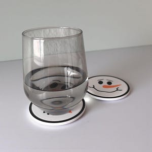 May include: A clear glass of water sits on a white coaster with a black border and a snowman face design. Another coaster with the same design is next to the glass. The coasters are round.