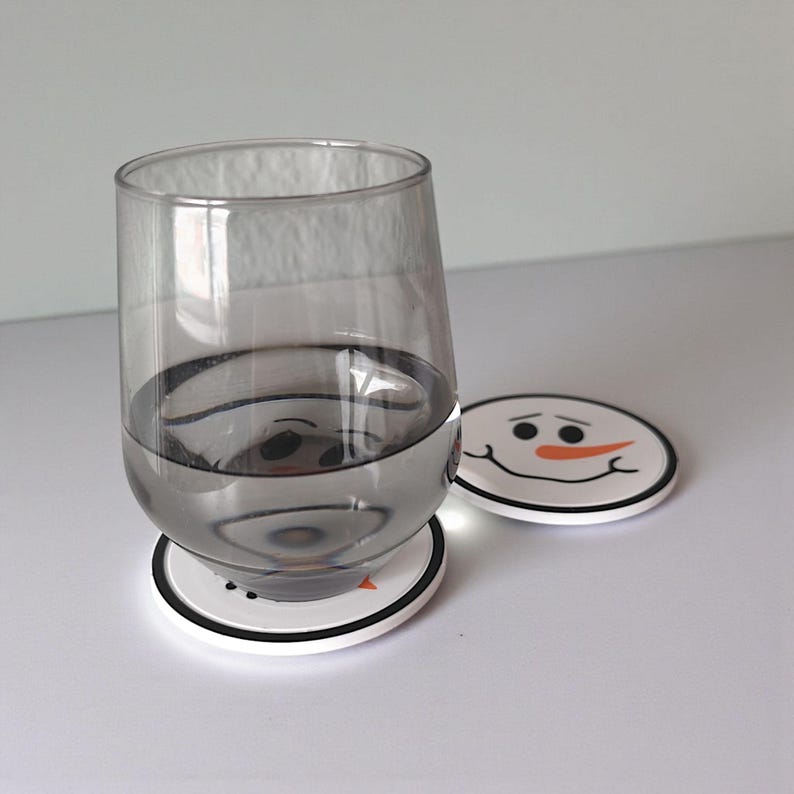 May include: A clear glass of water sits on a white coaster with a black border and a snowman face design. Another coaster with the same design is next to the glass. The coasters are round.