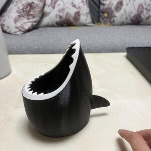 May include: A black and white shark-shaped container with a wide open mouth and a fin. The container has a white, jagged edge resembling teeth. The shark-themed item is sitting on a light-colored surface.
