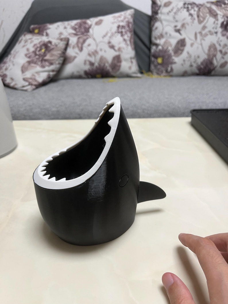 May include: A black and white shark-shaped container with a wide open mouth and a fin. The container has a white, jagged edge resembling teeth. The shark-themed item is sitting on a light-colored surface.